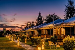 A row of covered outdoor seating areas with tables and chairs, surrounded by well-maintained lawns and trees, under a twilight sky. - Rawai Luxury Tents (Pushkar)
