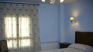A bedroom with two beds, a chair, a nightstand, a wall-mounted lamp, and a window with floral curtains. - Casa Rural Las Murallas (Brihuega)