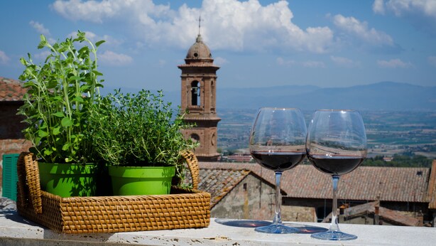 A terrace with a wicker basket containing green plants, two wine glasses with red wine, and a view of a bell tower and distant landscape. - Dimora Dell'Erbe (Montepulciano)