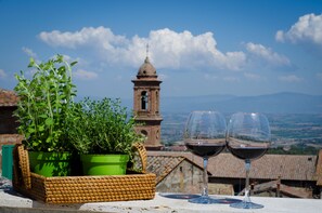 Una terraza con una cesta de mimbre que contiene plantas verdes, dos copas de vino tinto y vistas a una torre campanario y un paisaje lejano.