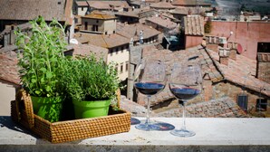 A balcony with a wicker basket containing green plants, two wine glasses with red wine, and a view of a town with terracotta rooftops. - Dimora Dell'Erbe (Montepulciano)