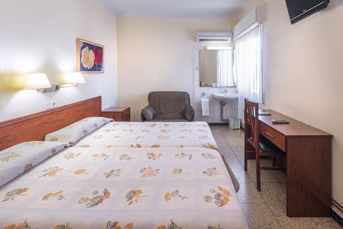 A hotel room with two beds, a desk, a chair, a bathroom with a sink and mirror, and an air conditioning unit.