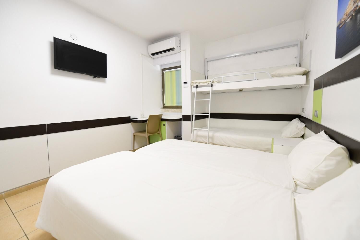A hotel room with a bunk bed, a desk with a chair, a flat-screen TV, and an air conditioning unit.