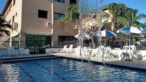 A swimming pool with a fountain, surrounded by white lounge chairs and umbrellas, with a building in the background. - Bella Casa Flat Service (Bertioga)
