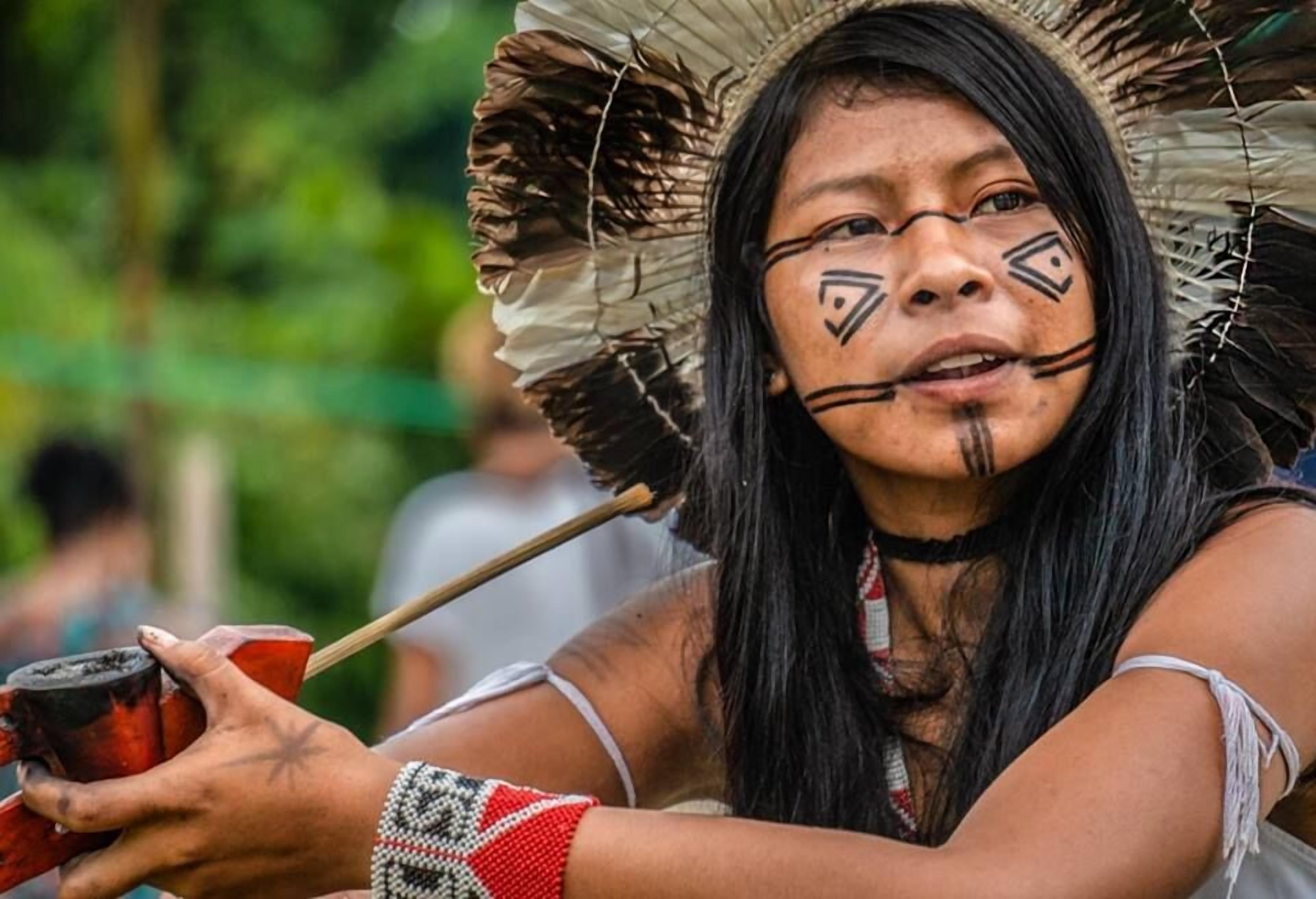 Una persona sosteniendo un objeto tradicional, vistiendo una prenda estampada y un tocado emplumado.