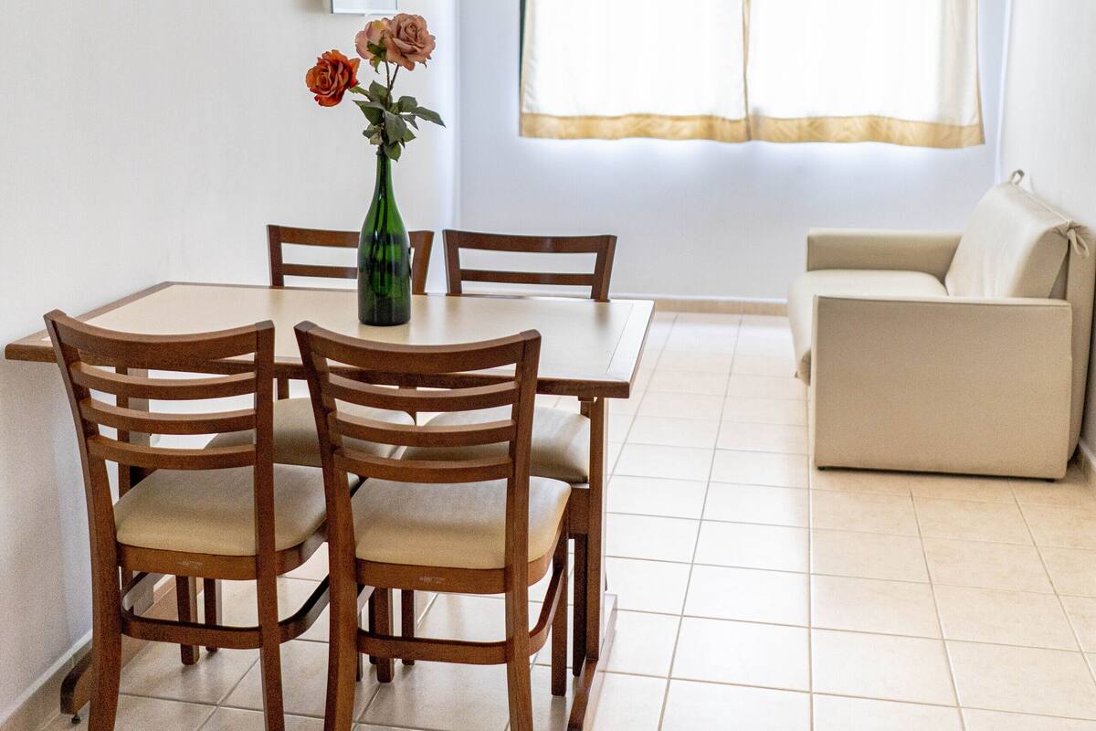 A dining area with a table and chairs, a vase with flowers, a ceiling fan, and a window with curtains.