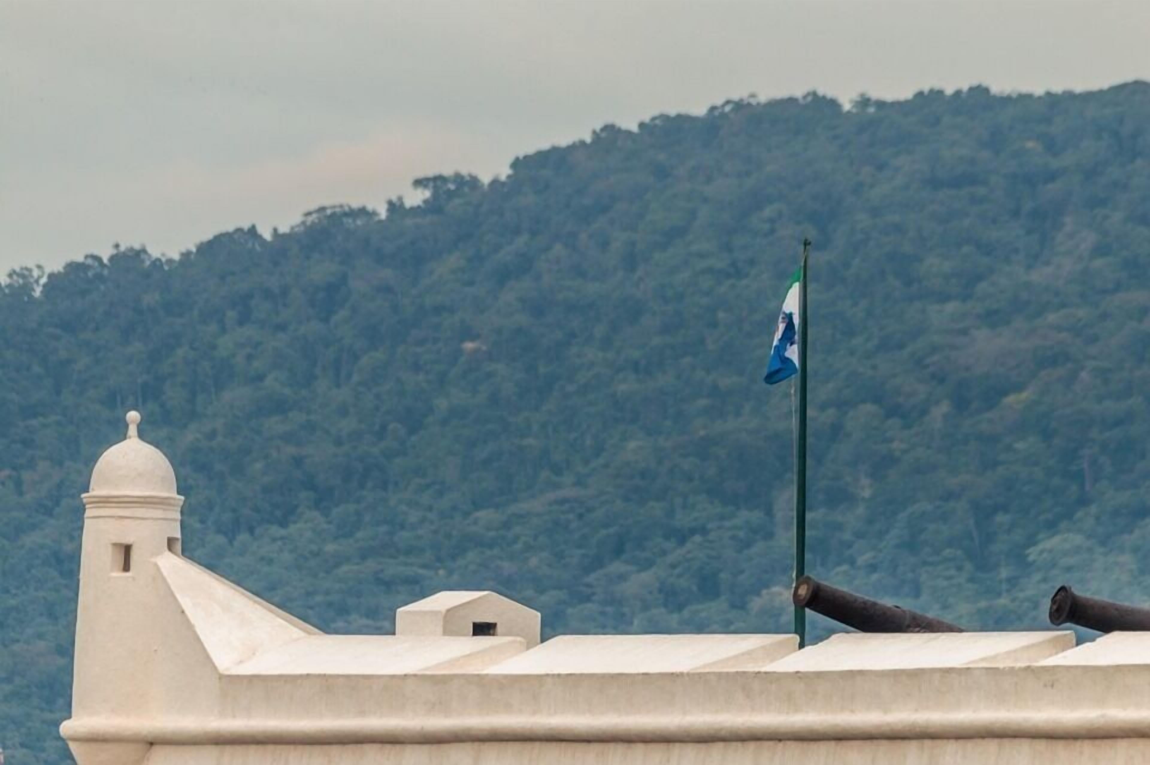 A building with a white exterior and a flagpole with a flag. There is a hill with trees in the background.