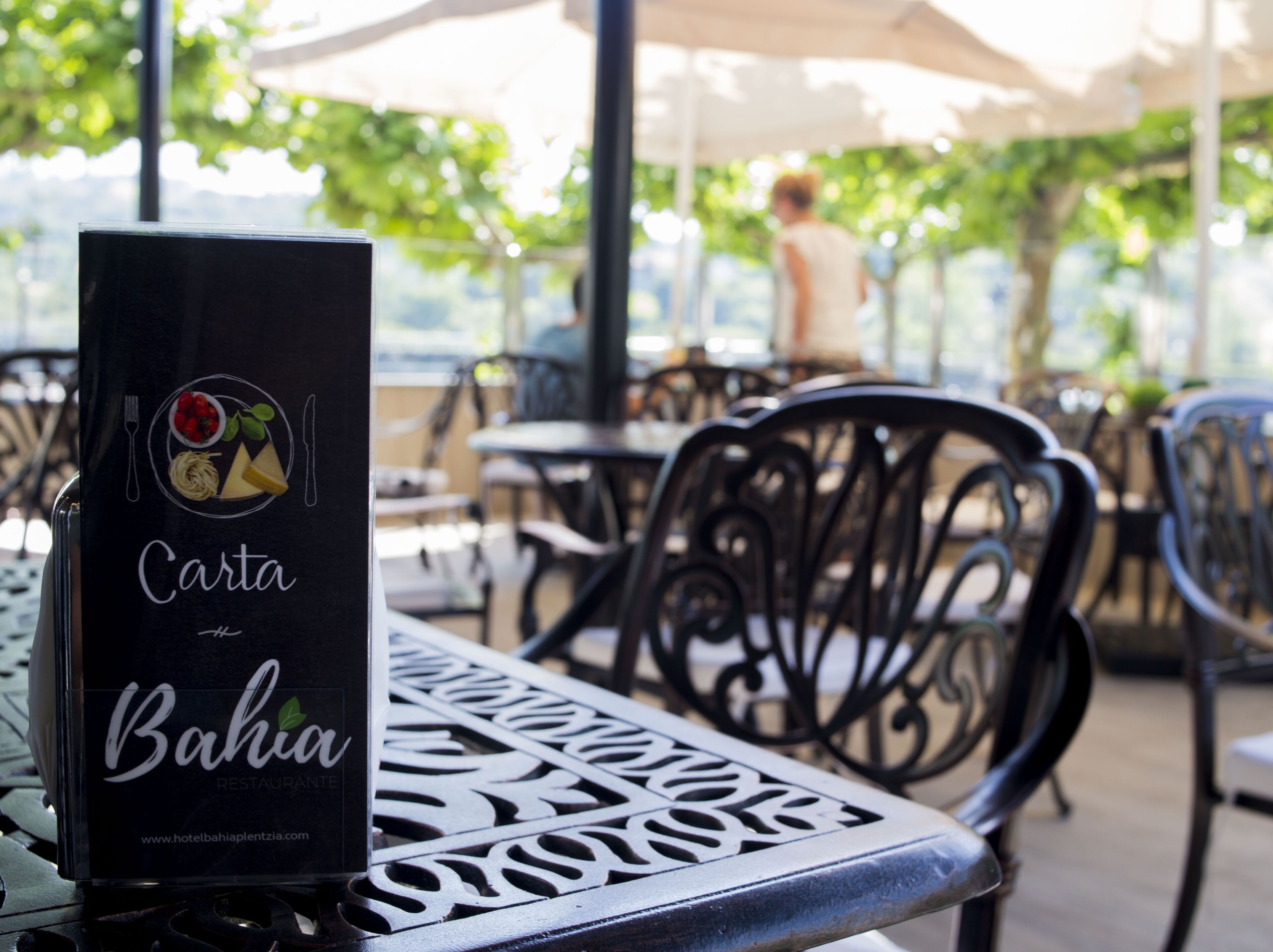 A menu stand with a logo and text for a Bahia restaurant, set on an outdoor table with metal chairs and umbrellas in the background.
