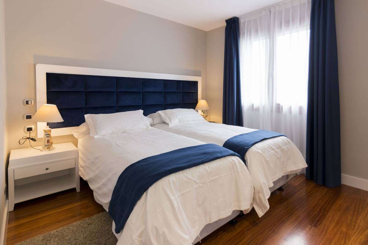 Cama doble con cabecera oscura, ropa de cama blanca y manta azul. Hay una mesita de noche con una lámpara y una pequeña mesa con un reloj. El cuarto tiene piso de madera y una ventana con cortinas azules.