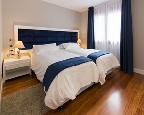 A double bed with a dark headboard, white bedding, and a blue throw blanket. There is a nightstand with a lamp and a small table with a clock. The room has wooden flooring and a window with blue curtains. - Hotel Boutique Bahía de Plentzia (Plencia)