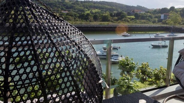 A balcony with a hanging chair, a table, and a view of a river with boats. - Hotel Boutique Bahía de Plentzia (Plencia)