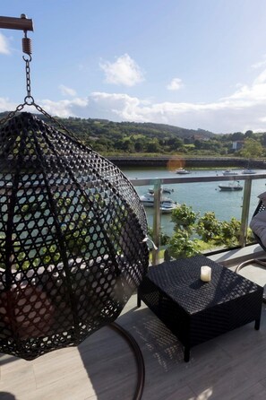 Un balcon avec un fauteuil suspendu, une table et une vue sur une rivière où naviguent des bateaux.