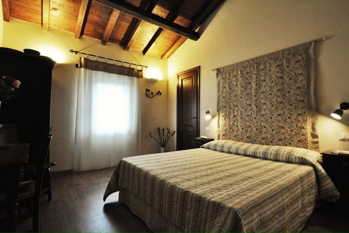 Une chambre à coucher avec un lit, des poutres en bois au plafond, une fenêtre avec des rideaux, une table de chevet et une lampe fixée au mur.