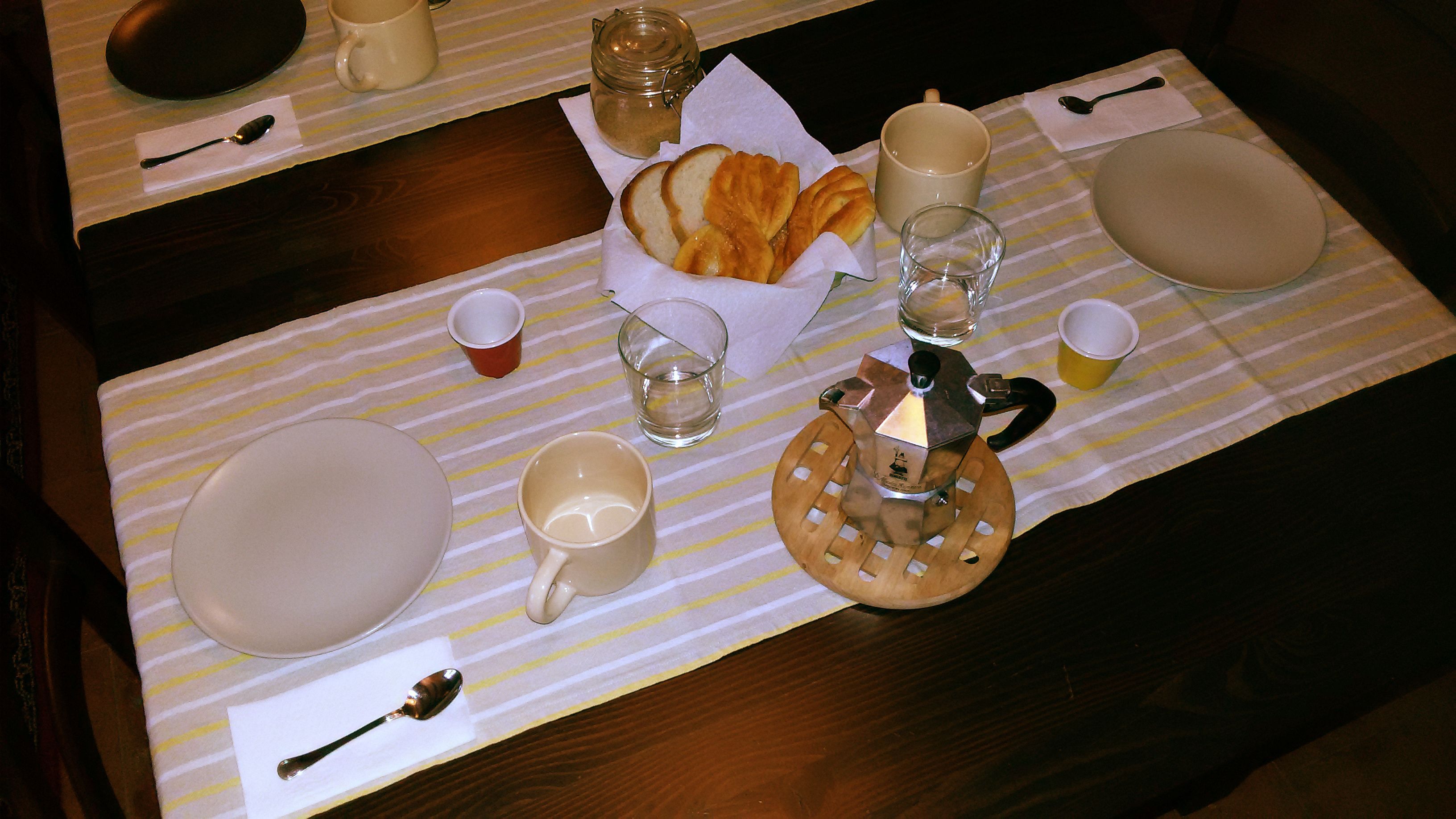 A breakfast setting with a coffee pot, cups, plates, and a pastry basket on a striped tablecloth.