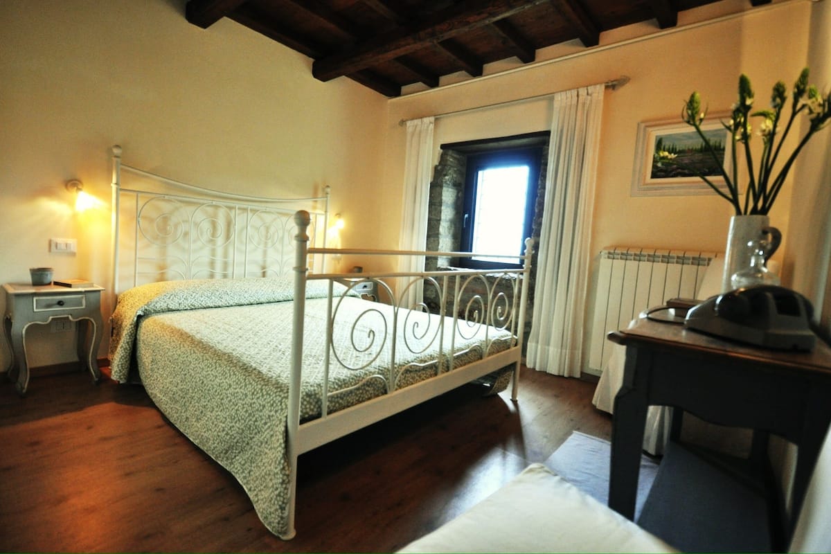 Une chambre à coucher avec un lit à ossature métallique, une table de chevet, une commode, une fenêtre avec des rideaux et un radiateur.