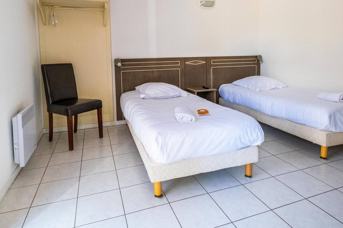 Habitación de hotel con dos camas individuales, una silla, una mesita y una lámpara en la pared.