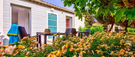 A white building with a covered patio area, outdoor seating, and a garden with yellow flowers.