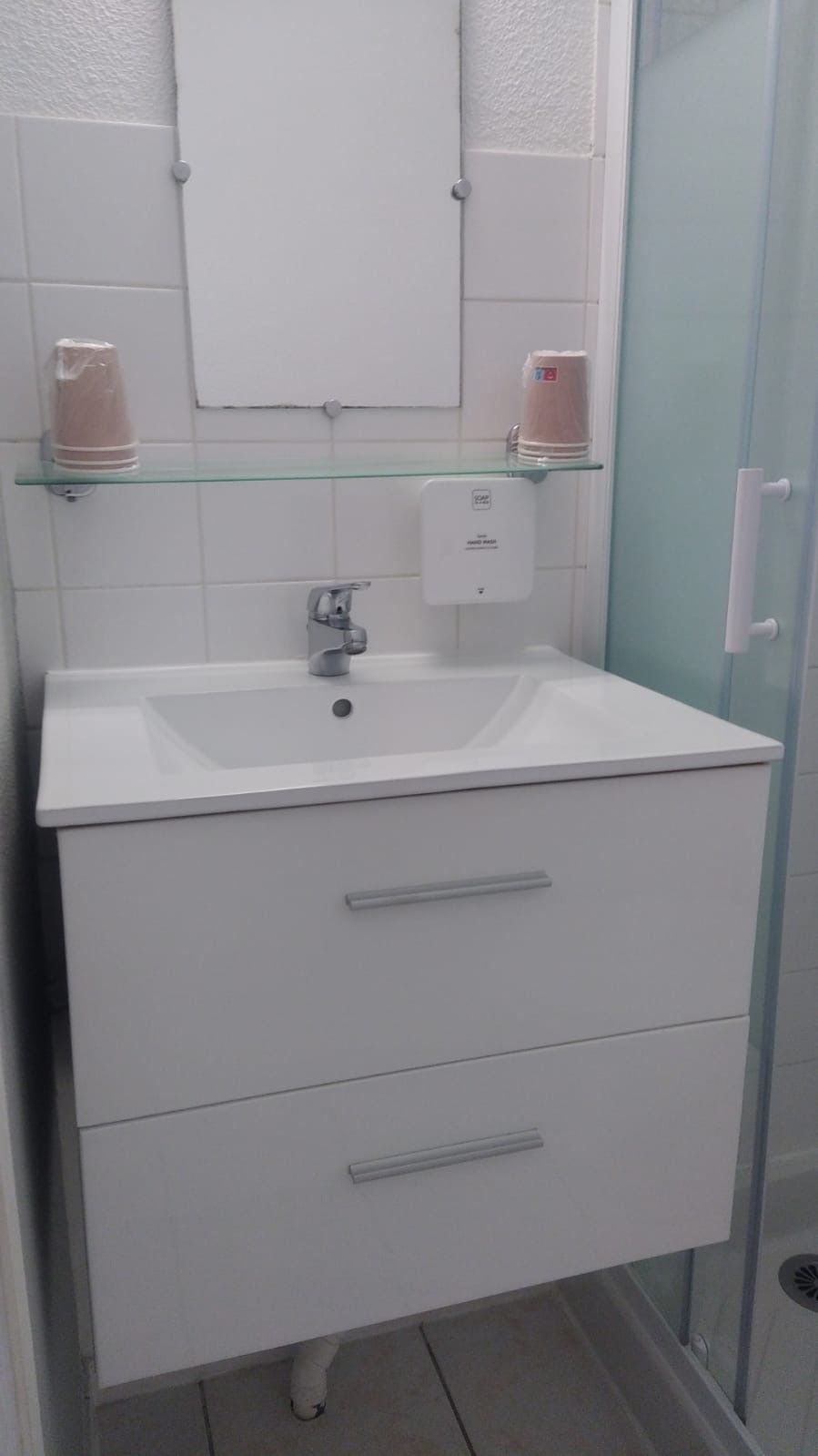 A bathroom with a white sink, two drawers, a glass shelf with toiletries, and a glass shower enclosure.