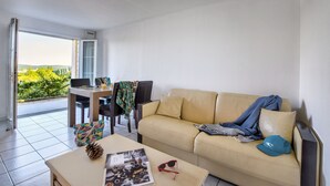 A living room with a beige sofa, a coffee table, and a dining table with chairs. There is a view of greenery through a sliding glass door. - Olydea Les Epesses (Les Epesses)