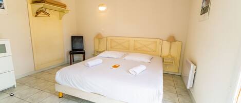 A neatly made bed with white linens and towels, a bedside table with a lamp, and a wall-mounted shelf with folded towels.
