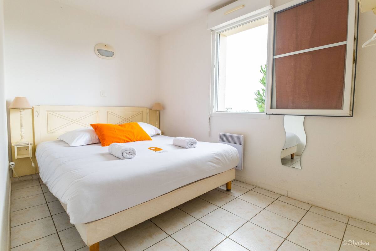 Una cama bien tendida con ropa de cama blanca y naranja, una lámpara de noche y una ventana con vistas a la vegetación.