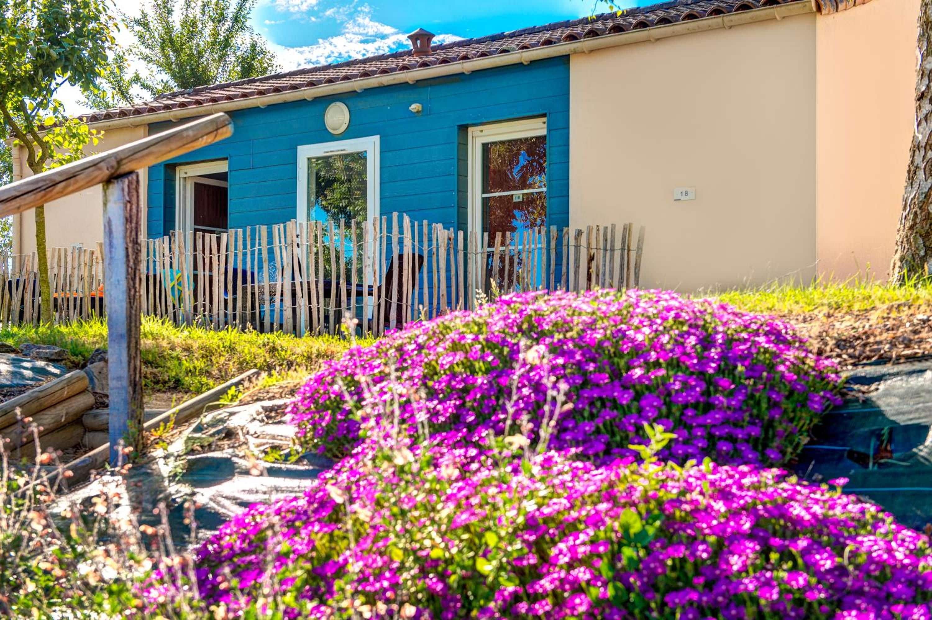 Una casa con fachada azul, una valla de madera y un jardín con flores moradas.