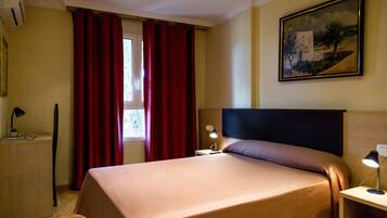 Una habitación de hotel con una cama, una mesita de noche, una lámpara, una ventana con cortinas rojas y un cuadro en la pared.