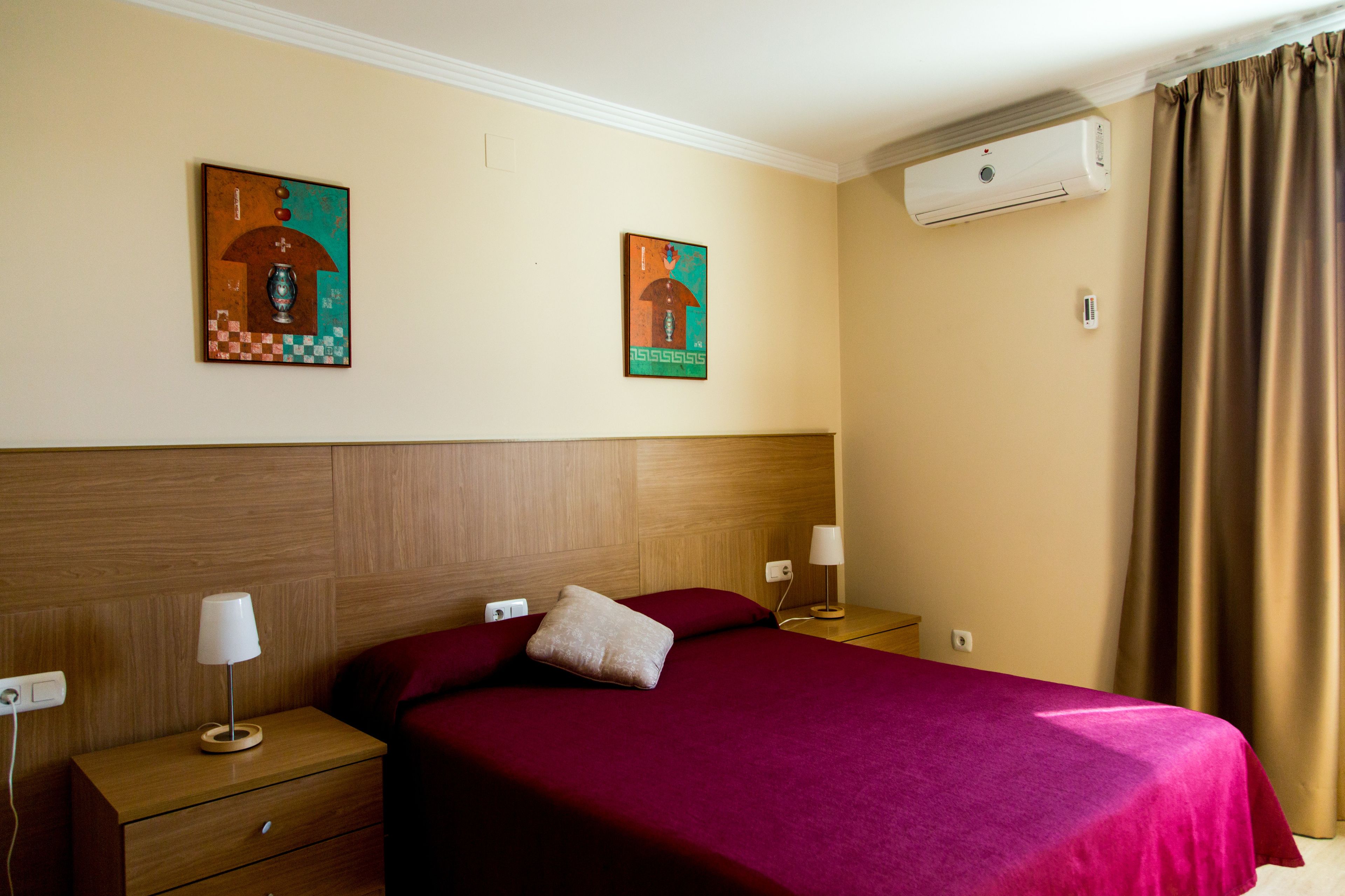 Habitación de hotel con cabecera de madera, cama grande con colcha roja, dos mesitas de noche con lámparas y dos unidades de aire acondicionado fijadas a la pared.
