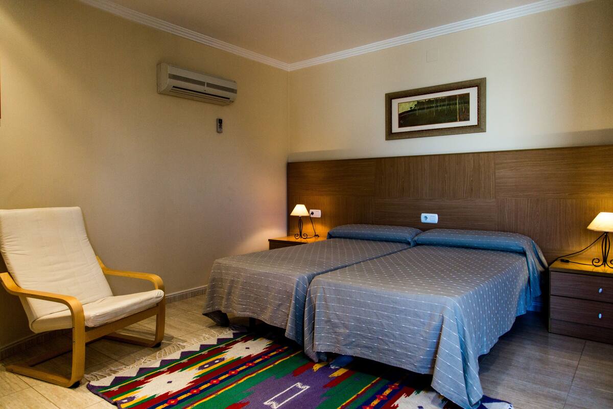 Quarto de hotel com duas camas, uma cadeira, uma mesinha de cabeceira e ar condicionado fixado na parede.