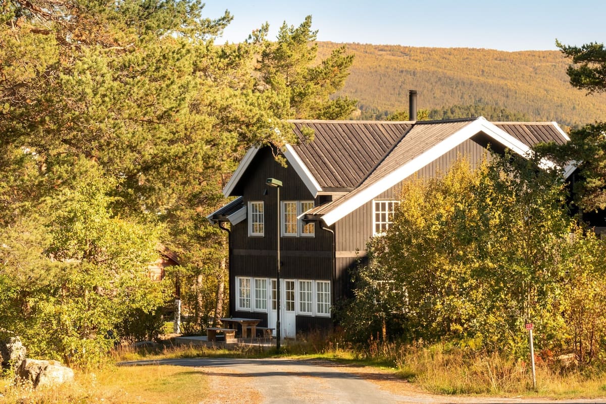 A two-story wooden house with a sloped roof, surrounded by dense trees and a mountain in the background.