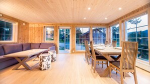 Un intérieur moderne avec des planchers et un plafond en bois, une table à manger avec des chaises, un canapé et de grandes fenêtres offrant une vue sur l’extérieur.