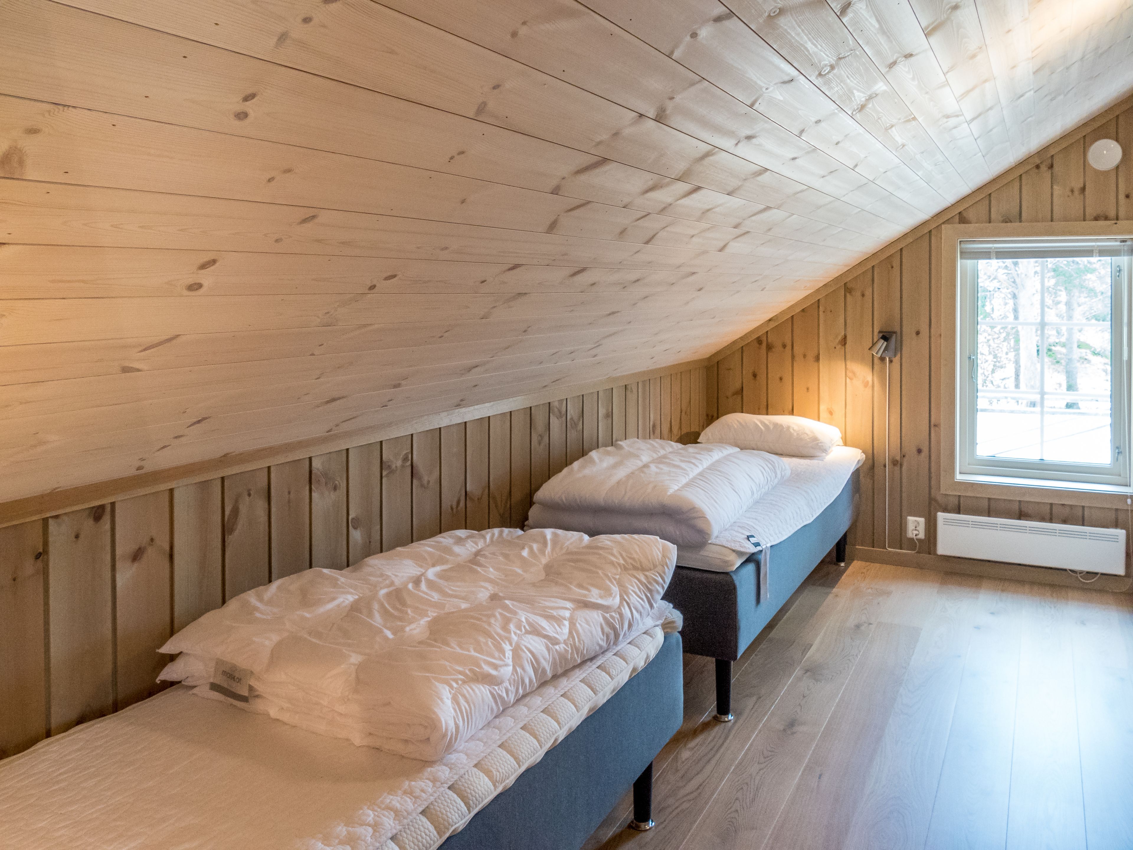 A room with wooden walls and a sloped ceiling, featuring two beds with white bedding and a window providing natural light.