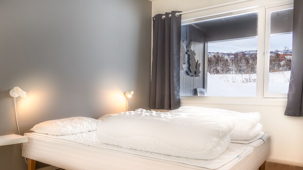 Un dormitorio con cama, lámparas de noche, ventana con cortinas y vistas a un paisaje nevado.