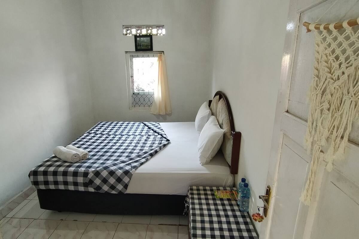 A small, simple room with a bed, a window with a curtain, a small table with a checkered tablecloth, and a macrame wall hanging.