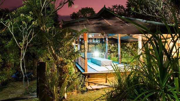 Un espace extérieur abrité, doté d’une terrasse en bois et d’un toit en métal, et entouré d’une végétation luxuriante.
