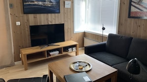 A modern living room with a wooden TV stand, a flat-screen TV, a dark sofa, a coffee table with a tray, and a window with blinds. - Slåttastølen Leiligheter by Geilolia (Hol)