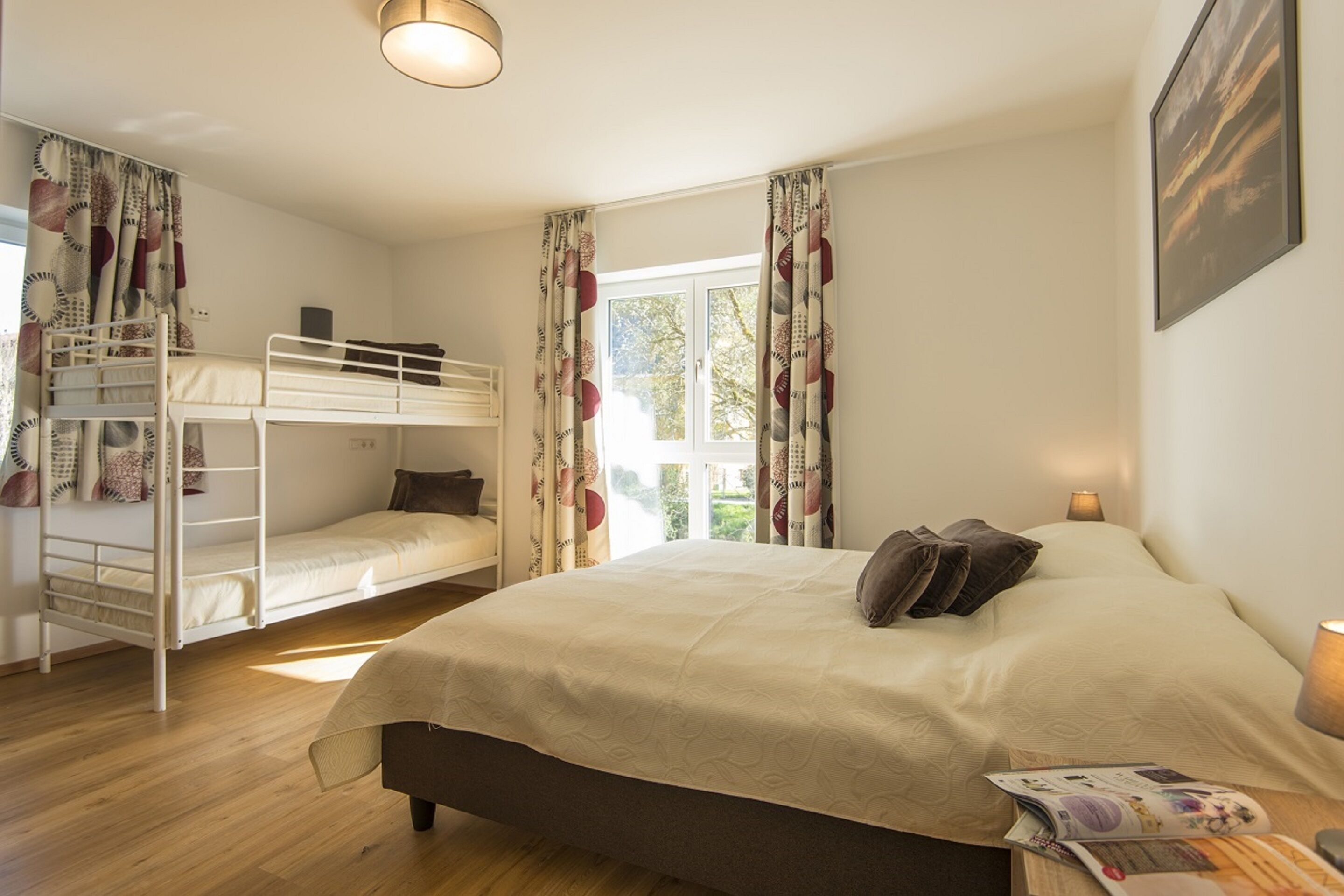 Une chambre avec un grand lit, un lit superposé, une fenêtre avec des rideaux, une lampe et un tableau encadré au mur.