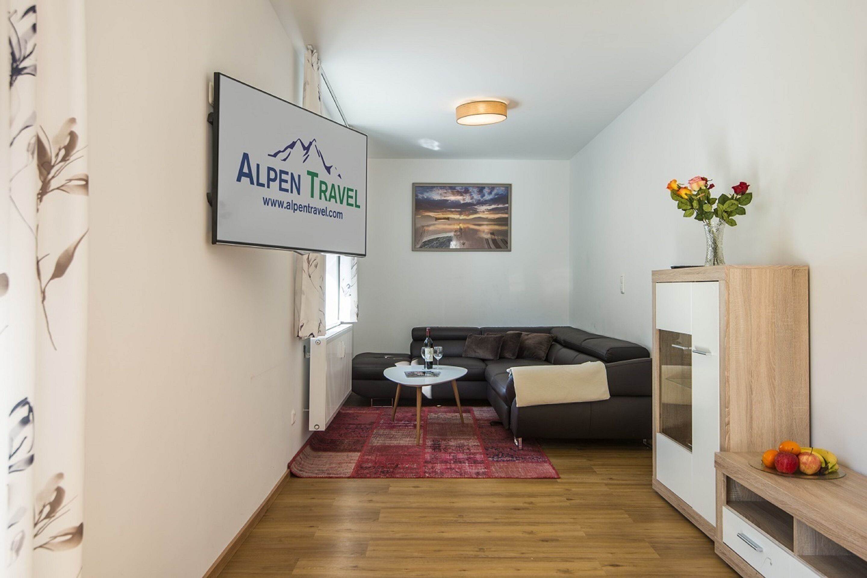A modern living room with a sofa, a small round table with a bottle and glass, a red rug, a framed picture on the wall, and a cabinet with fruit on top.