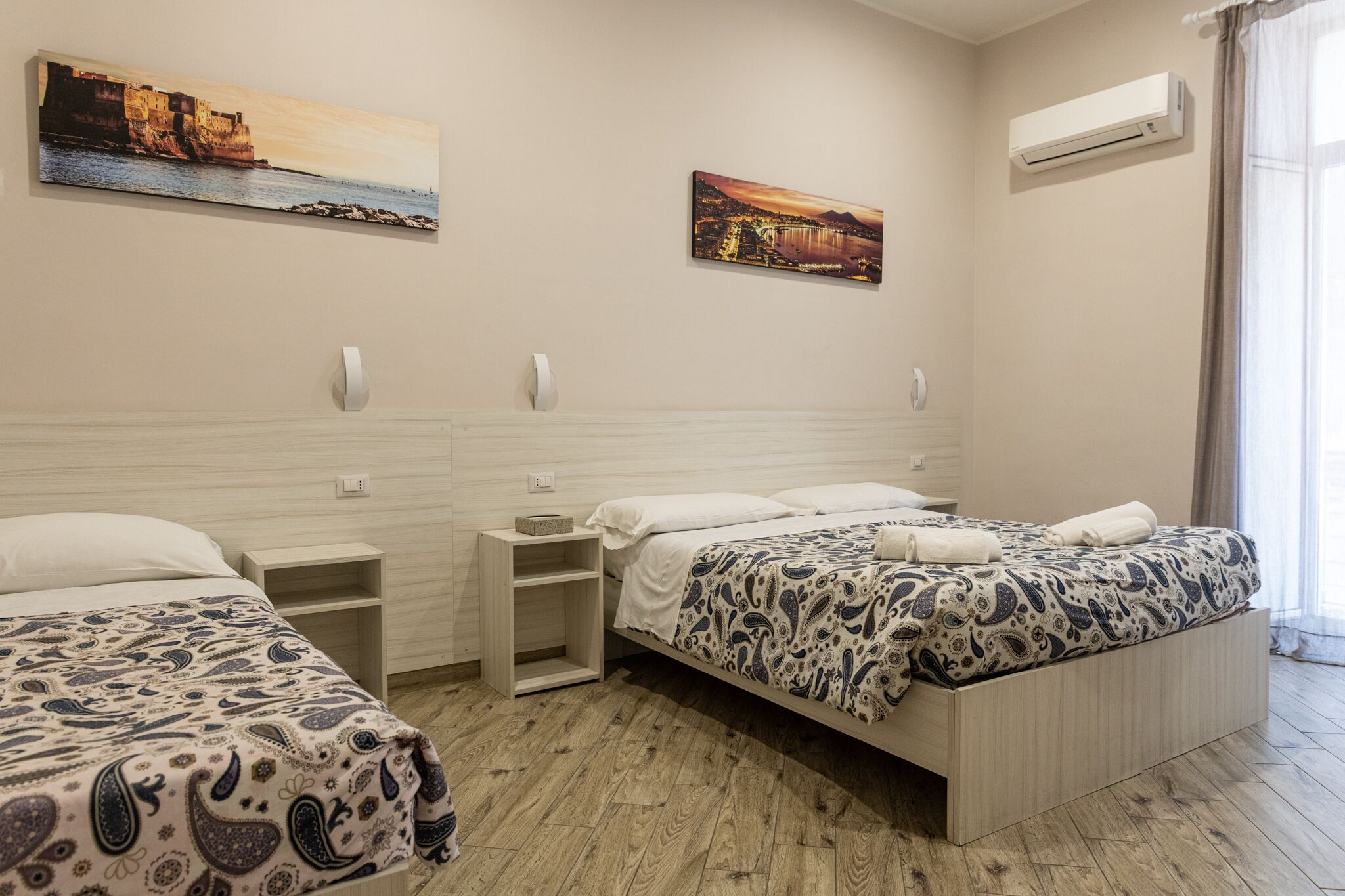 A hotel room with two beds, a wooden headboard, a nightstand, and a wall-mounted air conditioner.