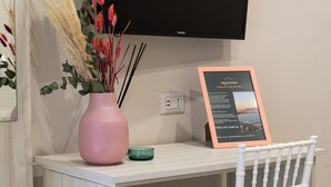 A hotel room with a white desk, a pink vase with dried flowers, a framed welcome sign, a television mounted on the wall, and a white chair. - B&B alla Ferrovia (Naples)