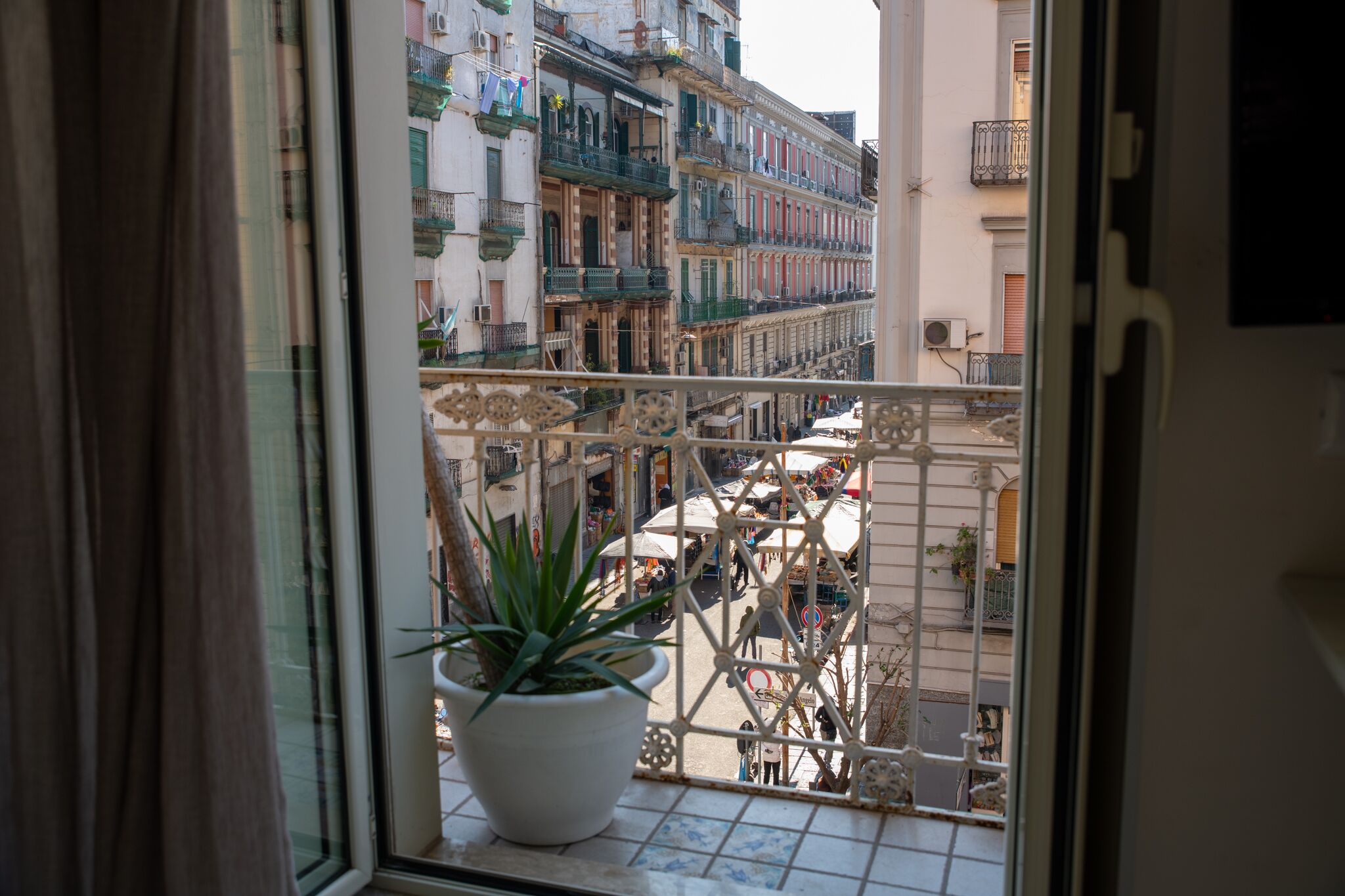 A balcony with a potted plant, overlooking a busy street with buildings and shops. - B&B alla Ferrovia (Naples)