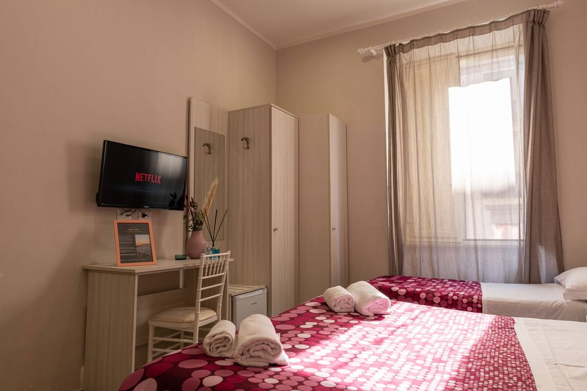 Habitación de hotel con una televisión mostrando 'NETFLIX', un escritorio con una silla, dos camas con ropa de cama roja y blanca, un armario y una ventana con cortinas traslúcidas.