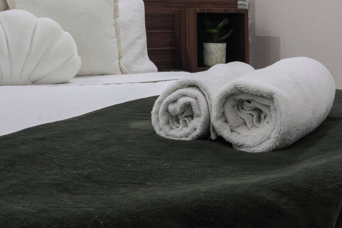 A neatly made bed with white linens and a dark green blanket, a wooden headboard, and a wall with a leaf-patterned wallpaper.