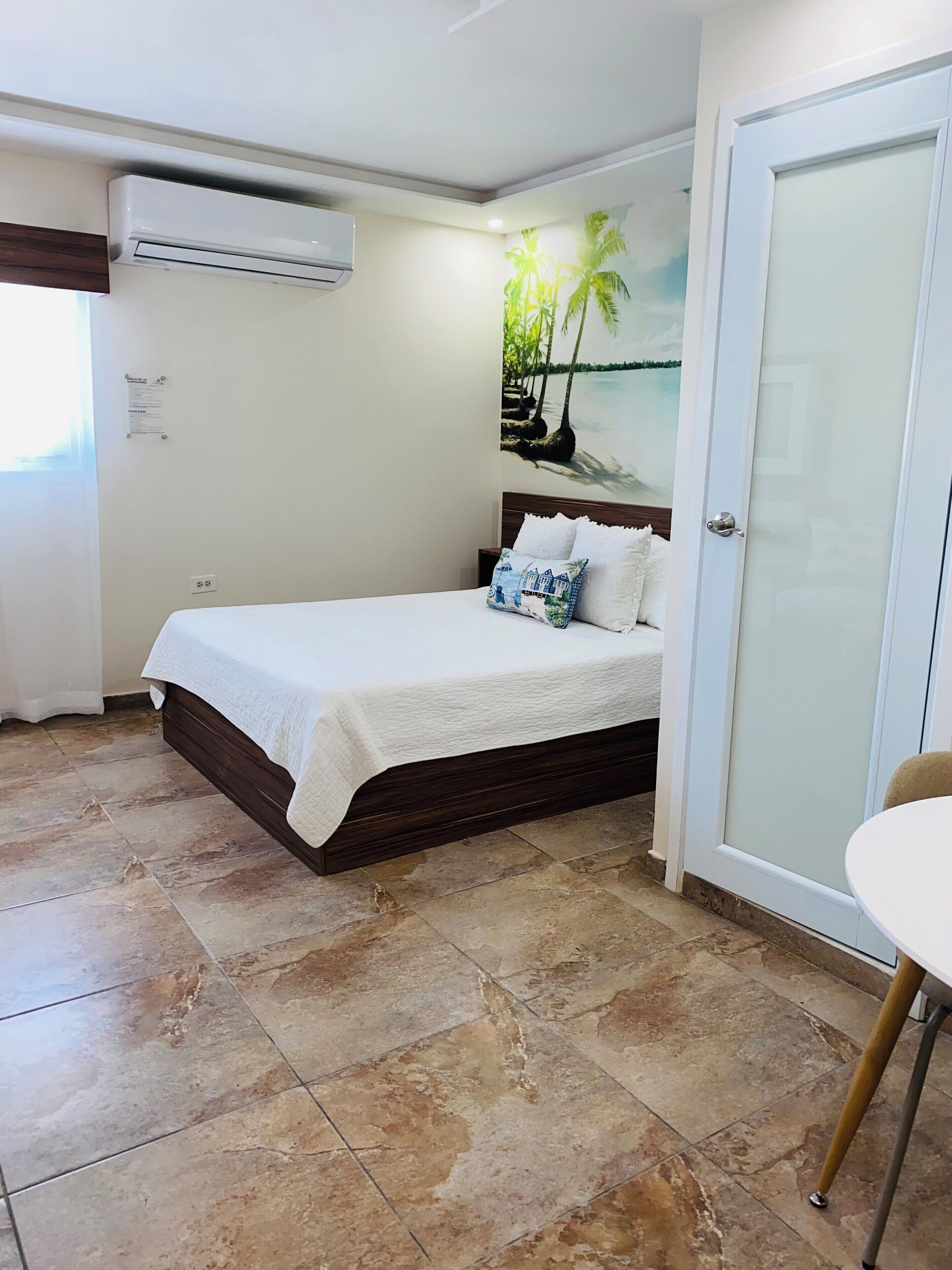 A bedroom with a bed, an air conditioning unit, a door, and a wall with a tropical mural.