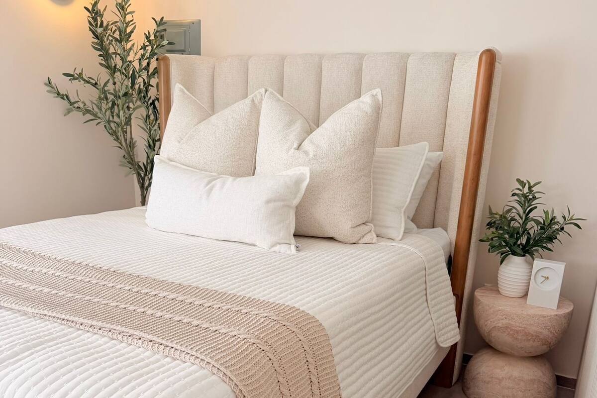 A neatly made bed with a headboard, a bedside plant, and a wall-mounted light fixture.