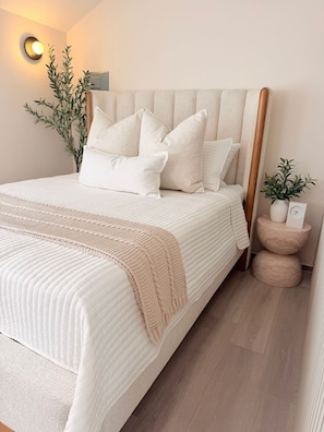 Una cama bien hecha con cabecera, una planta en la mesita de noche y una lámpara fijada a la pared.
