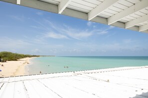 Vista a la playa desde un área techada con techo blanco, cielo azul claro y mar turquesa.