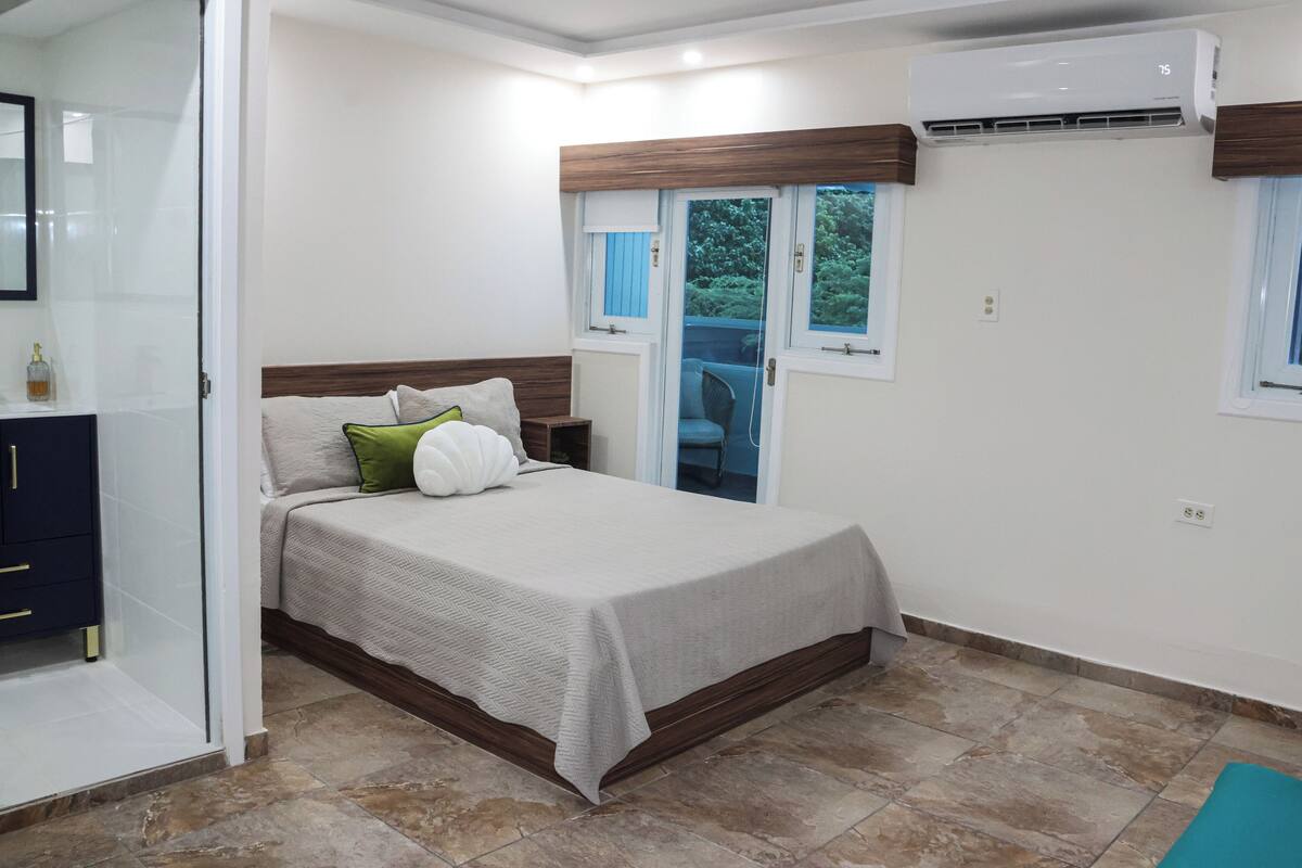 A bedroom with a bed, a window, an air conditioning unit, and a bathroom with a shower.