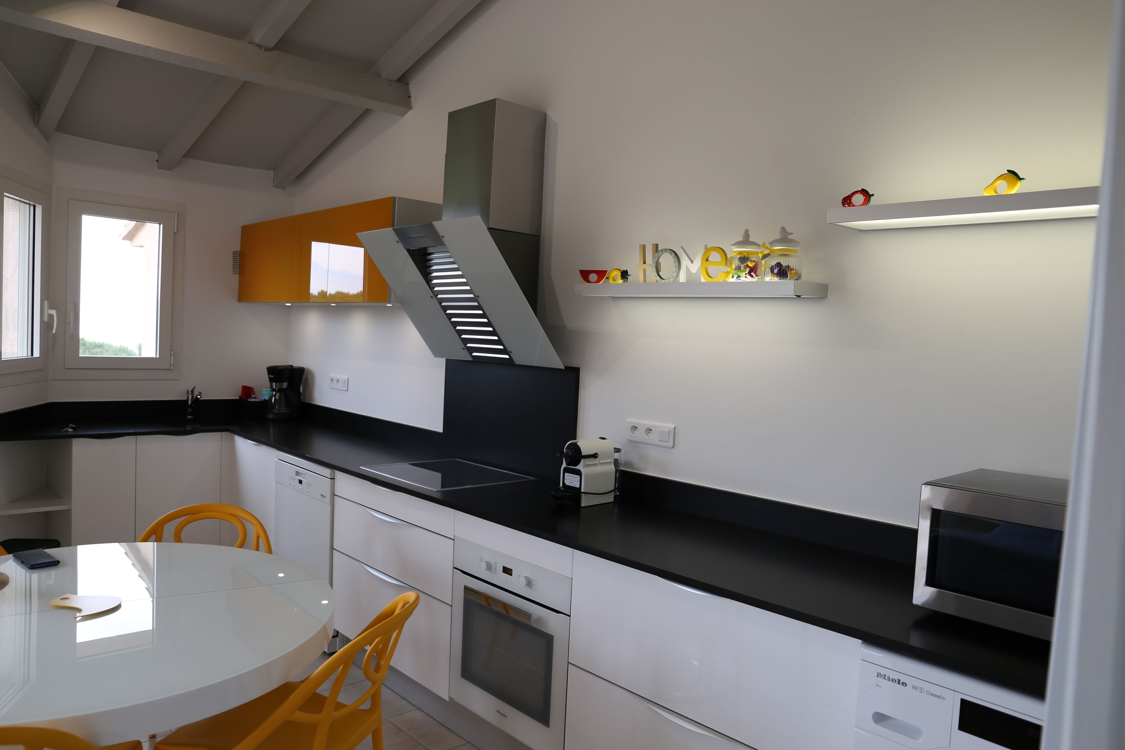 Una cocina moderna con armarios blancos, encimera negra y horno empotrado. Hay una ventana que proporciona luz natural y una estantería con objetos decorativos.