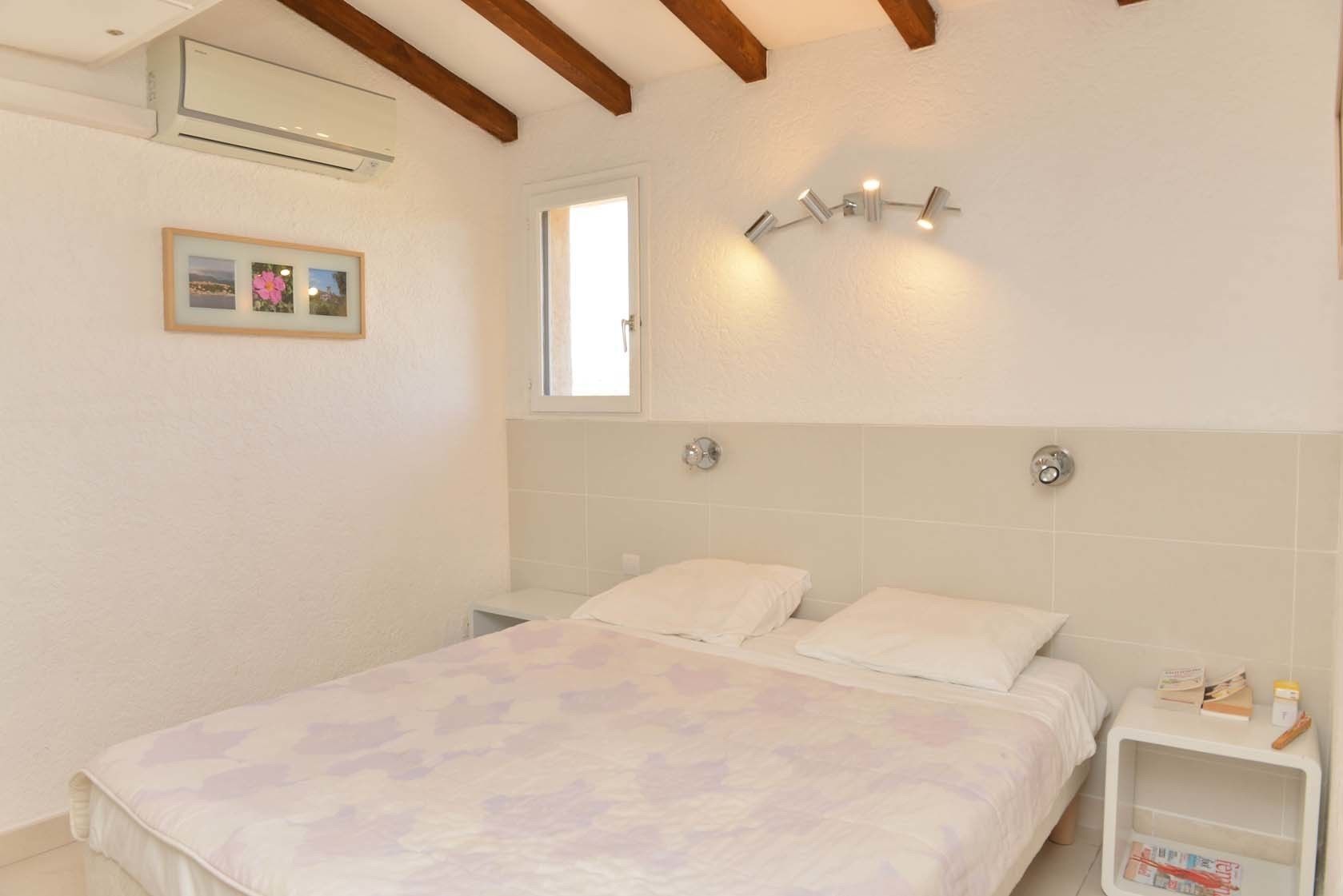 A bedroom with a bed, a bedside table, a nightstand, a window, and a wall-mounted light fixture.